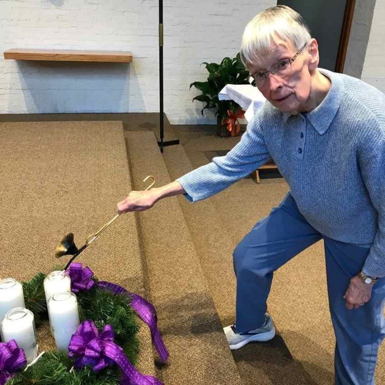 An older woman with short gray hair leans down to light the first candle of an Advent wreath.