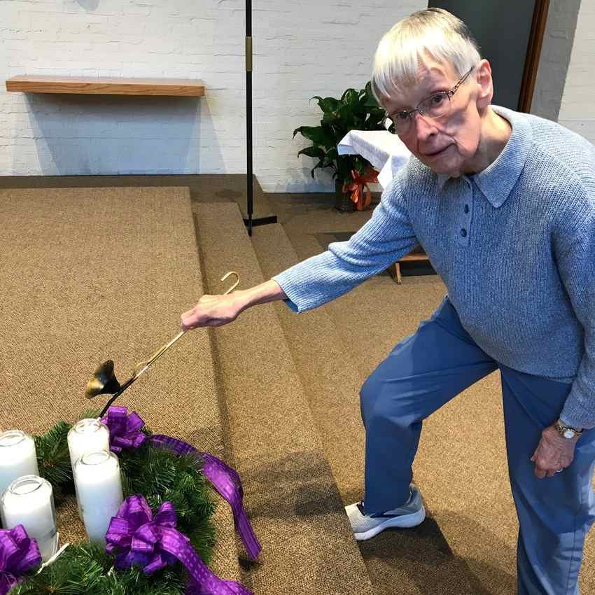 An older woman with short gray hair leans down to light the first candle of an Advent wreath.