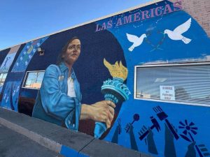 A mural with a hispanic woman holding a statue of libery tourch looking into the distance. The top of the mural has doves coming together and a sign that says "Las Americas"