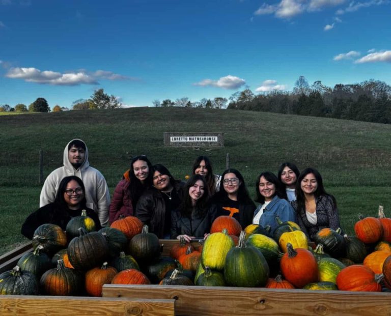 A group of young Latino students standing outside behind a cart of colorful pumpkins. Behind them is a hill with a stone white sign that says "Loretto Motherhouse"