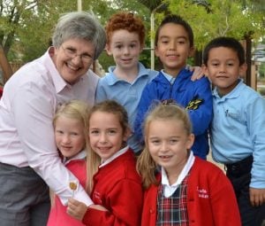 A woman hugs a group of elementary children wearing school uniforms.