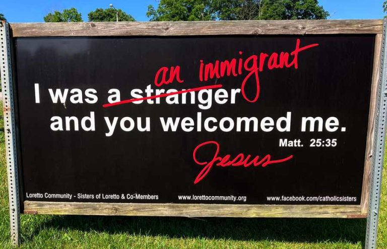 Large black sign with "I was a stranger and you welcomed me. Matt. 25:35" in white text, with "a stranger" crossed out with red and "an immigrant" written over it in red. "Jesus" is signed in red.