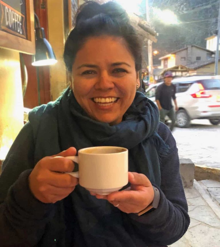 A Latina woman in her late 30ss early 40s smiles holding a cup of coffee outside a coffee shop. She is wearing a blue sweater and scarf and her black hair is in a high ponytail.