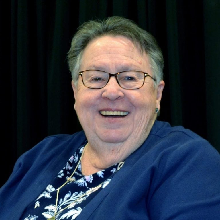 A headshot of an older woman smiling brightly. She has short grey and brown hair, rectangular tortoise glasses and is wearing a blue shirt.