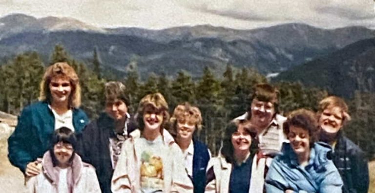 A blurry older photo of a group of adults and kids in front of Colorado mountains smiling for a photo on a hike.