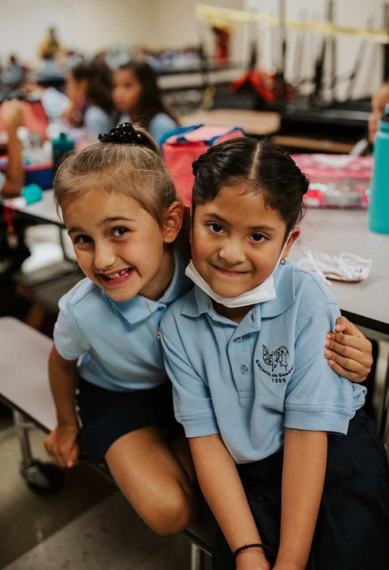 Two young elementary aged Latina girls smiling together on the first day of school. Their uniforms are blue shirts with "Escuela de Guadalupe" printed on them.