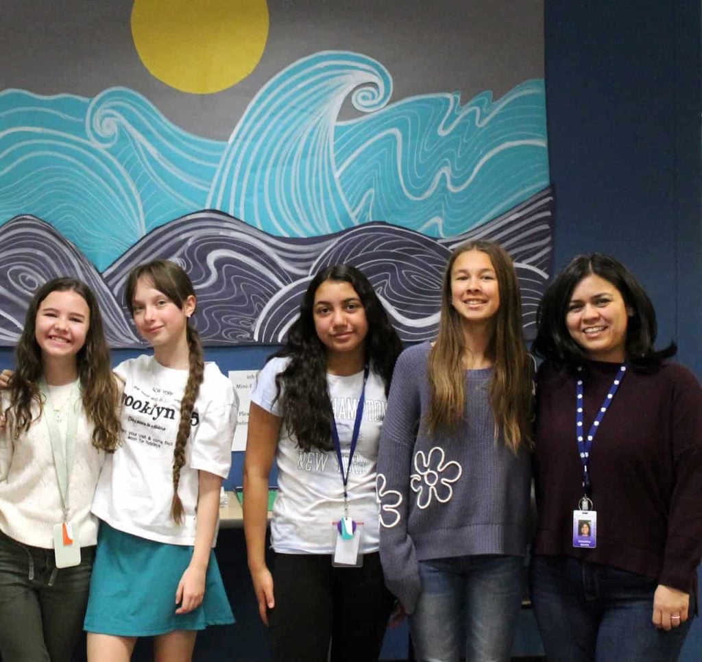 A teacher taking a photo with four of her middle school advisory group students in front of a mural of abstract waves.