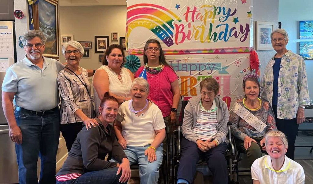 A group of staff and residents at a group home smiling together in front of a Happy Birthday sign for one of the resident's birthdays.