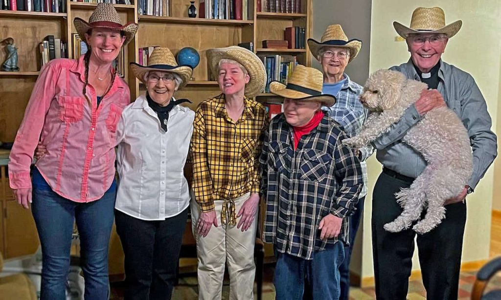 A group of older folks, including a man holding a white fluffy dog, are smiling and wearing straw cowboy hats.