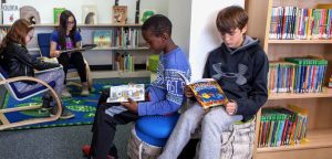 Four elementary aged students sitting on stools and chairs reading books in a school library.