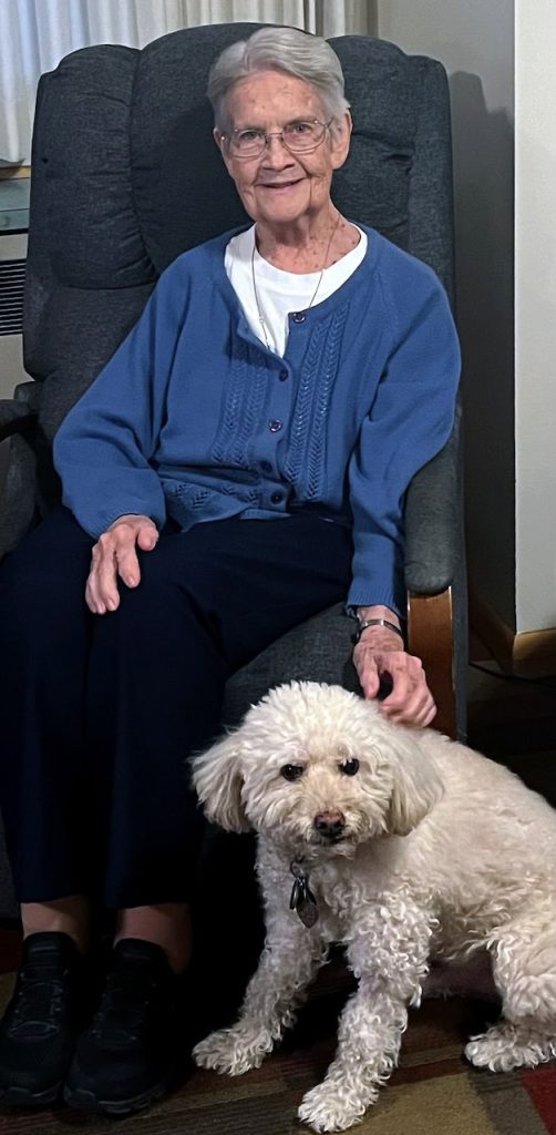 An older white woman with grey hair, glasses and a blue cardigan relaxes in a comfy chair as she pets a white fluffy dog by her side.
