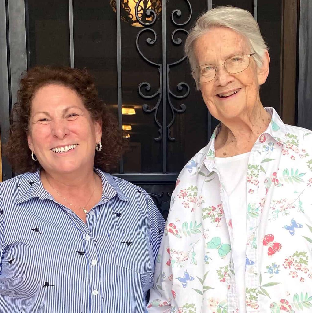 A short woman with curly brown hair, silver hoop earrings and a blue and white vertical striped t-shirt is standing next to an older woman with white hair, glasses and a floral t-shirt. They are smiling.
