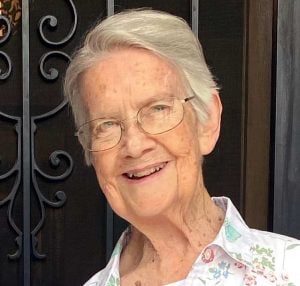 An older white woman wearing glasses smiles for a photo outside a decorative metal door.