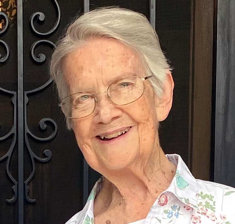 An older white woman wearing glasses smiles for a photo outside a decorative metal door.
