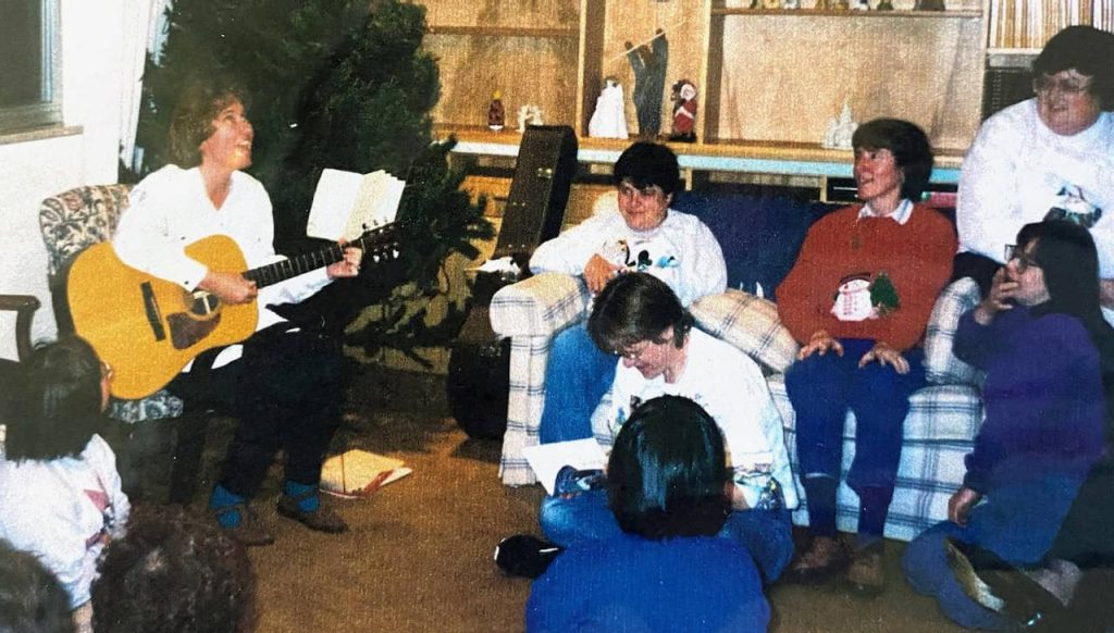 A joyous holiday time with people in a community sitting on the floor and a couch smiling and listening to a woman playing guitar and singing.
