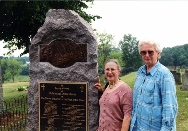 Two white women standing next to a memorial for enslaved persons on the Loretto Motherhouse campus in Nerinx, Kentucky. The plaque reads "In loving memory of oblates and other slaves of the Sisters of Loretto at the foot of the cross."