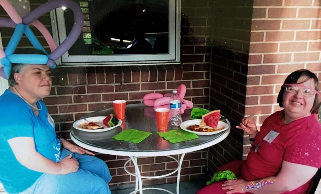 Two women with disabilities eating lunch together. The woman on the left has a balloon had on and is looking into the distance. The woman on the right is smiling at the camera and has glasses and a red shirt.