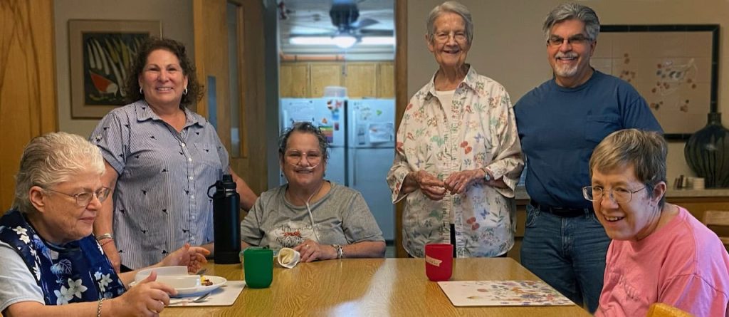 A group of people in a community living house sitting and standing around a table smiling.