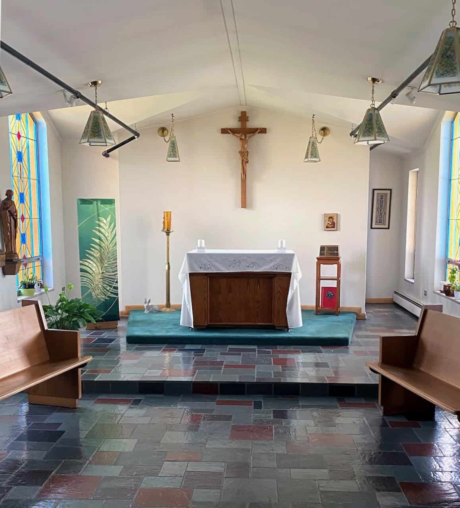 A small chapel with stained glass window panes, a couple of wooden pews on either side of an altar.