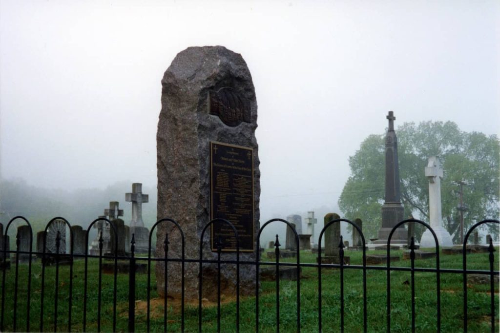 A large slab of stone with a plaque on it in the Loretto Motherhouse cemetery on a cloudy day. It is a memorial to those who were enslaved at the Motherhouse.