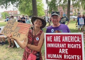 Two people pose for a photo with their signs at a protest in a park. The woman's sign reads "We are not ok" with an upside-down American flag. The man's sign reads "We are America: Immigrant rights are human rights!"