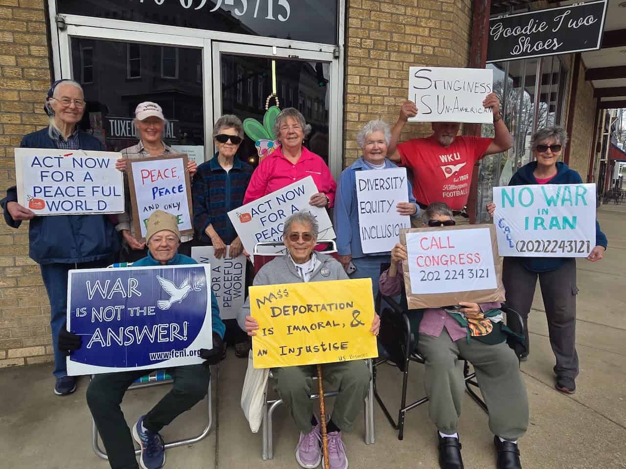 A group of older people holding signs of protest gather for a photo. Three are in chairs in the front; the other seven stand behind them.