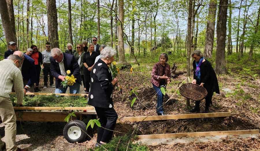 People gather around the grave in the woods to place greens and flowers into the grave.
