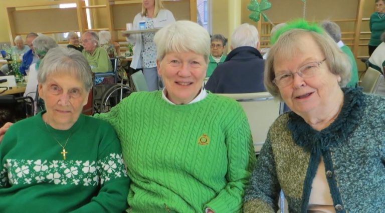 Three older women dressed festively in green and shamrocks lean in for a photo together in the Motherhouse dining room.