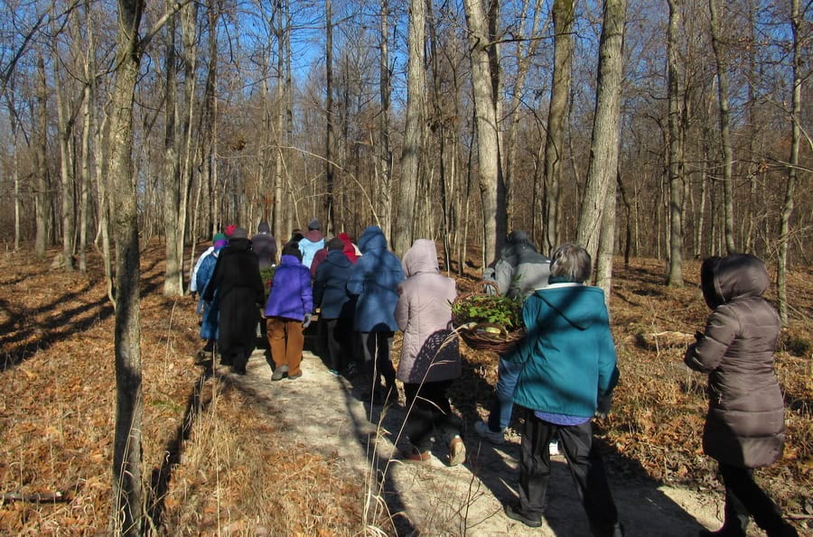 People dressed in winter coats processing along a path into a snowless winter woods.