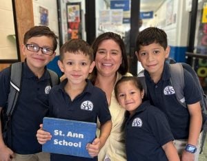 Four elementary students in school uniform polo shirts and big smiles gather round Kathleen Fox, principal at St. Ann's School, for a group photo.