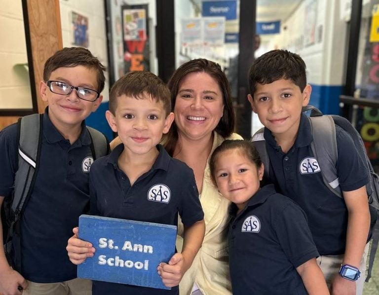 Four elementary students in school uniform polo shirts and big smiles gather round Kathleen Fox, principal at St. Ann's School, for a group photo.