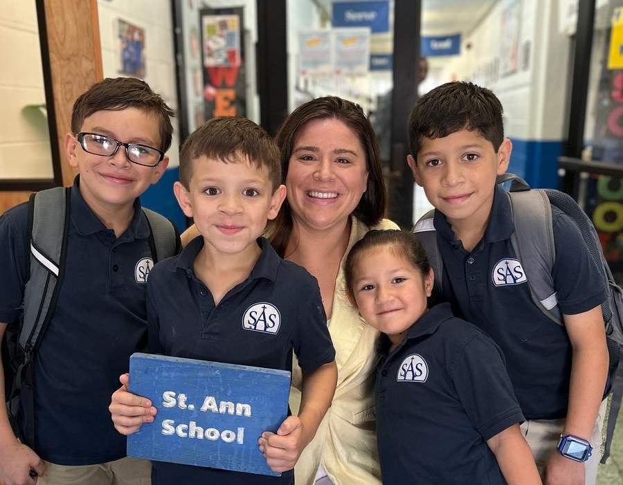 Four elementary students in school uniform polo shirts and big smiles gather round Kathleen Fox, principal at St. Ann's School, for a group photo.
