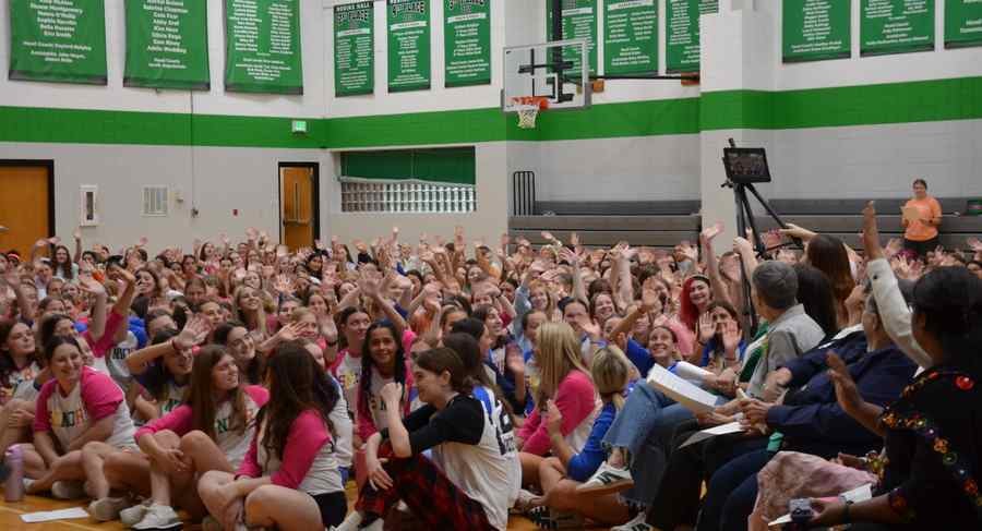 High school students sitting on the Nerinx Hall gym floor have turned to wave at people on the right side of the photo. The girls are waving at members of the Loretto Community present in the assembly.