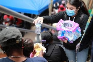 A young Latina in a mask reaches up the stairs to hand a bottle of water to a Black child eating on her caregivers lap.