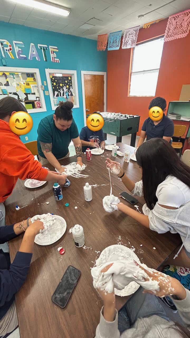 A volunteer at a table with a bunch of elementary aged kids shown making slime together.