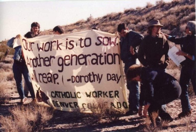 Photo from 1989 of eight people holding up a large fabric banner amongst the Nevada scrub desert. The banner reads "Our work is to sow. Another generation will reap. - Dorothy Day Catholic Worker, Denver, Colorado