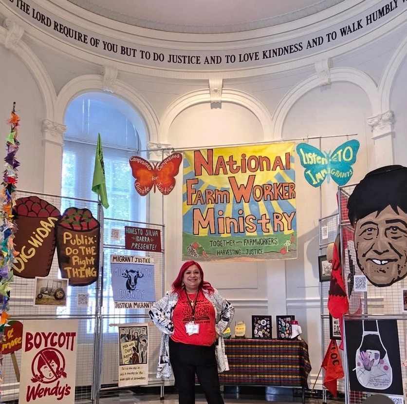 A Latina woman with dyed red hair and a bright smile stands with her fists on her hips in front of a National Farm Worker Ministry display. The display fills much of the space in the domed room, with protest posters, a poster reading "National Farm Worker Ministry" in the middle and a larger-than-life cut out of a Latino man's face.