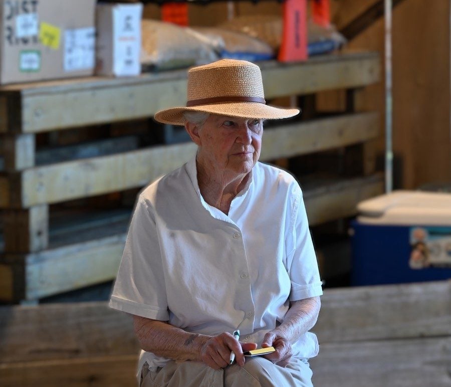 An older woman in a straw hat and holding a pen and reporter's note pad looks intently at something in the distance.