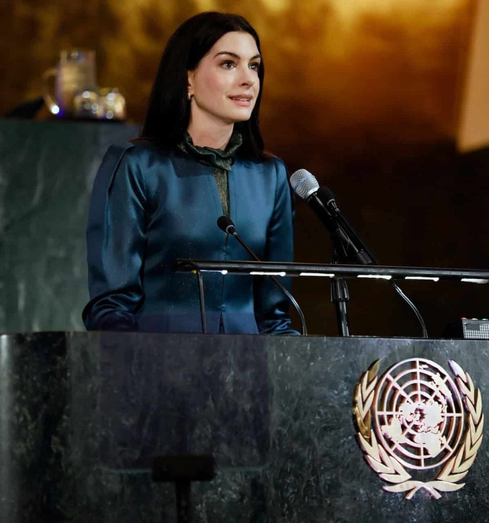 Anne Hathaway with shoulder length brown hair and a soft blue suit stands at the UN podium in the general hall and is giving a speech.