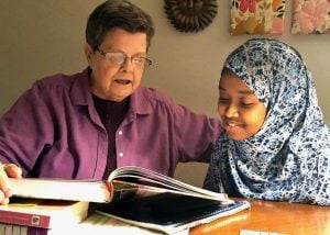 An older white woman with brown hair and glasses and a pink shirt is shown helping a young woman of color with her homework. She is wearing a beautiful blue and white patterned hijab.