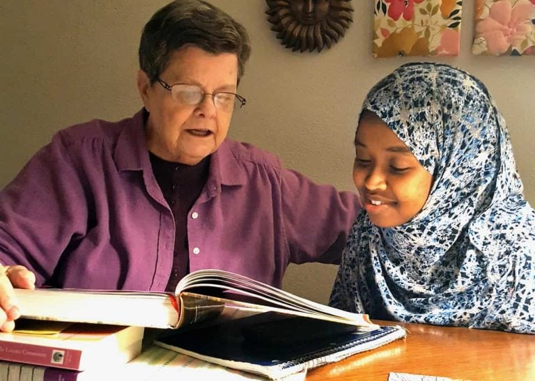 An older white woman with brown hair and glasses and a pink shirt is shown helping a young woman of color with her homework. She is wearing a beautiful blue and white patterned hijab.