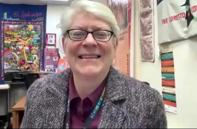 A middle-aged woman with short white hair and small rectangular glasses is smiling as she sits in an office decorated with bright colorful posters behind her.