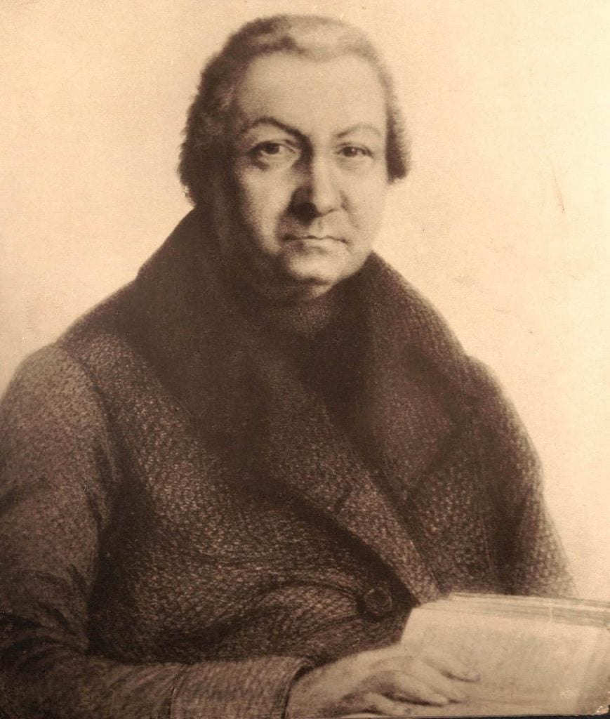 An archival black and white image of a man, Charles Nerinckx. He is wearing a tweed coat and his hand is on a open book.