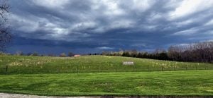 The Loretto Motherhouse entrance in Kentucky. A sprawling bright green grassy hill with a fence to keep in the cows and a sign that says "Loretto Motherhouse" in the distance. Half the frame are large pillowy clouds on a darker blue and grey day.