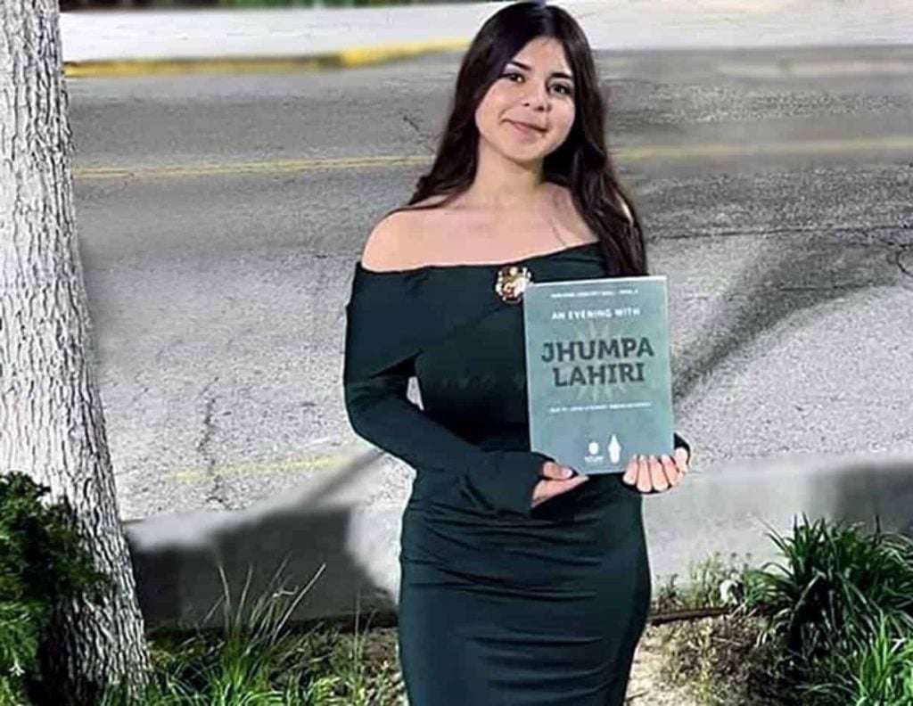A young Latina woman with long brown hair in a middle part with a semi formal fitted dark blue dress smiles softly as she holds a book that says "An Evening with Jhumpa Lahiri"