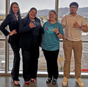 Four college students in business casual outfits standing together in front of a large window smiling at a conference.
