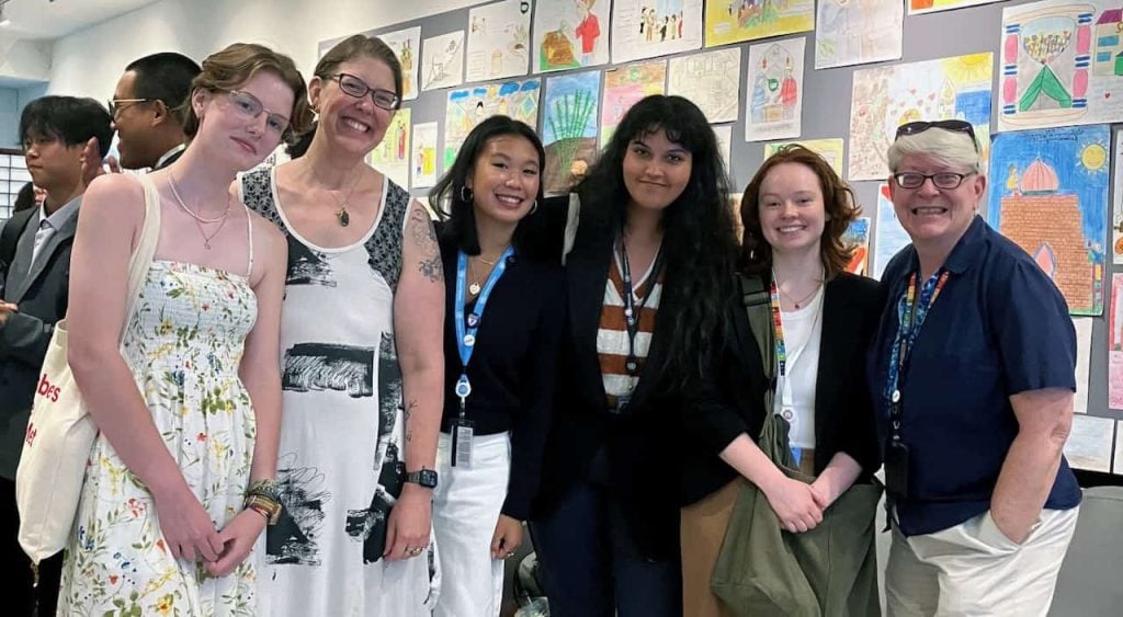 A six-person intergenerational group of femme people smiling together at a United Nations office.