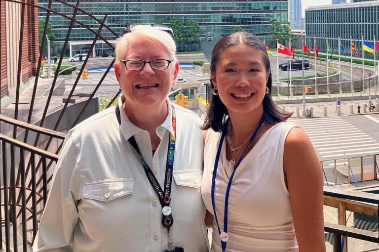 A middle aged woman with white hair, black glasses and a white polo smiles with a college aged women with shoulder length brown hair a white tank top and silver hoop earrings. They are in front of the United Nation. Behind them you can see an array of various nations flags.