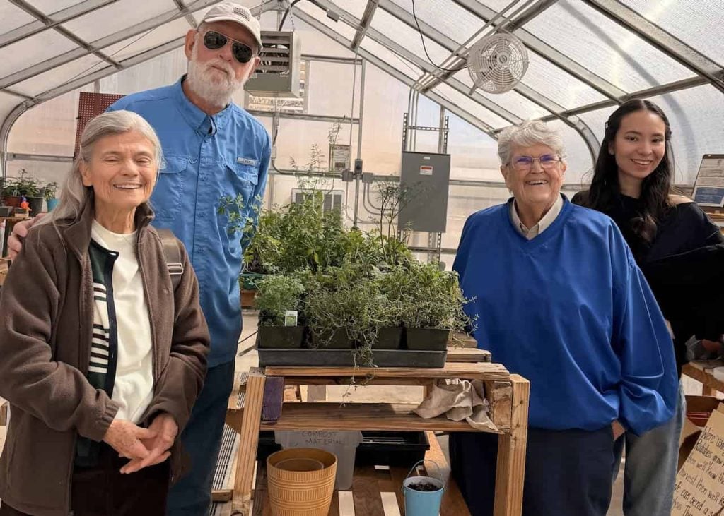 Three elders and a younger college student are pictures smiling together in a greenhouse with lots of green plants on a shelf in between them.