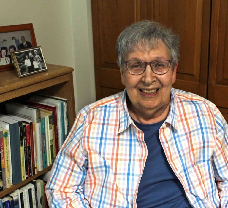 An older white woman sitting down smiling wearing glasses a blue t-shirt and a striped blue and orange collared long sleeve shirt over the blue shirt. She looks relaxed and is sitting next to her books on a bookshelf and on top of the shelf are family photos.